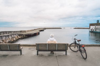 A young Caucasian woman sits with her back on a wooden bench overlooking the Baltic Sea on the seafront in Copenhagen Denmark in winter in cloudy weather. Girl walking gonoskoy bike parked nearby clipart