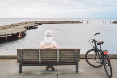 Genç bir beyaz kadın sırtında bulutlu havalarda kışın sahil Kopenhag Danimarka Baltık Denizi bakan bir tahta bankta oturur. Kız gonoskoy bisiklet yürüyüş yakın park
