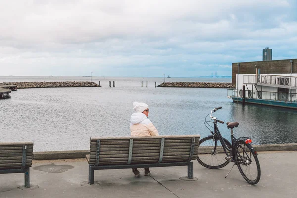 Seorang wanita muda Kaukasia duduk dengan punggungnya di bangku kayu yang menghadap Laut Baltik di tepi laut di Kopenhagen Denmark pada musim dingin dalam cuaca mendung. Gadis berjalan motor gonoskoy diparkir di dekatnya — Stok Foto