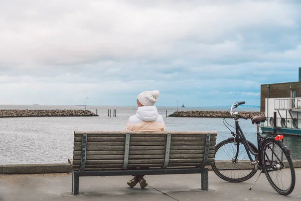 Молодая кавказская женщина сидит спиной на деревянной скамейке с видом на Балтийское море на берегу моря в Копенгагене зимой в облачную погоду. Девушка идет гоноской велосипед припаркован неподалеку — стоковое фото