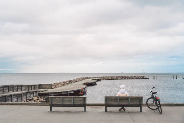 Uma jovem caucasiana senta-se de costas em um banco de madeira com vista para o Mar Báltico, em frente ao mar, em Copenhague, Dinamarca, no inverno, em clima nublado. Menina andando bicicleta gonoskoy estacionado nas proximidades — Fotos gratuitas