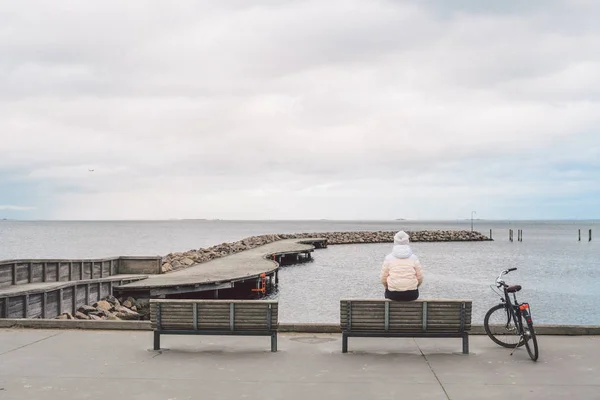 Молода жінка кавказьких сидить з її назад на дерев'яні лавки з видом на узбережжі Балтійського моря, на березі моря в Копенгагенській взимку в похмуру погоду. Дівчина, що йде gonoskoy велосипед припарковані поблизу — стокове фото