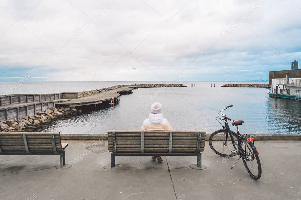 A young Caucasian woman sits with her back on a wooden bench overlooking the Baltic Sea on the seafront in Copenhagen Denmark in winter in cloudy weather. Girl walking gonoskoy bike parked nearby