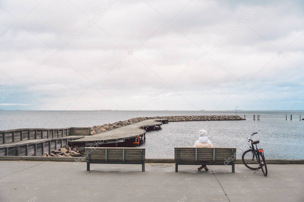 A young Caucasian woman sits with her back on a wooden bench overlooking the Baltic Sea on the seafront in Copenhagen Denmark in winter in cloudy weather. Girl walking gonoskoy bike parked nearby