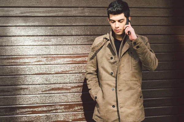 Portrait of a model young mulatto male Turkish Mediterranean race brunet in coat uses hand phone technology businessman on the street in sunny weather against the background of a wooden wall — Stock Photo, Image