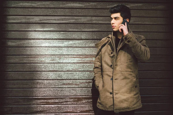 Портрет модели молодого мулата мужчины турецкой средиземноморской расы брюнет в пальто использует ручной телефон бизнесмен на улице в солнечную погоду на фоне деревянной стены — стоковое фото