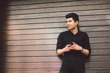 Genç yakışıklı Türk erkek manken portresi. Akdeniz yarış esmer siyah gömlekli bir ahşap duvar arka planda güneşli havalarda sokakta bir el telefon teknolojisi işadamı kullanıyor