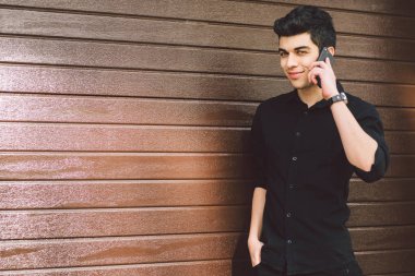 Portre modeli genç yakışıklı erkek şüpheli Türk Orta Doğu esmer siyah gömlekli bir ahşap duvar arka planı güneşli havalarda sokakta teknoloji çağırmak için el telefon kullanır