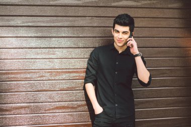 Portre modeli genç yakışıklı erkek şüpheli Türk Orta Doğu esmer siyah gömlekli bir ahşap duvar arka planı güneşli havalarda sokakta teknoloji çağırmak için el telefon kullanır