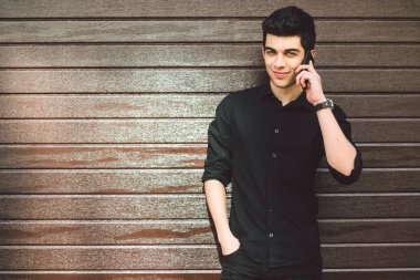 Portre modeli genç yakışıklı erkek şüpheli Türk Orta Doğu esmer siyah gömlekli bir ahşap duvar arka planı güneşli havalarda sokakta teknoloji çağırmak için el telefon kullanır