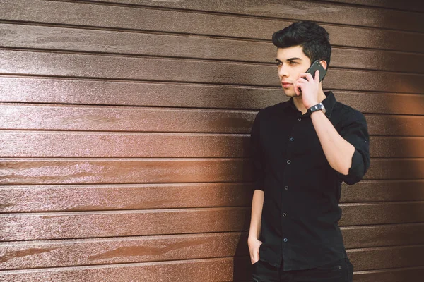 Modelo de retrato jovem bonito masculino suspeito turco médio oriental morena em camisa preta usa telefone de mão para chamar a tecnologia na rua em tempo ensolarado contra o fundo de uma parede de madeira — Fotografia de Stock