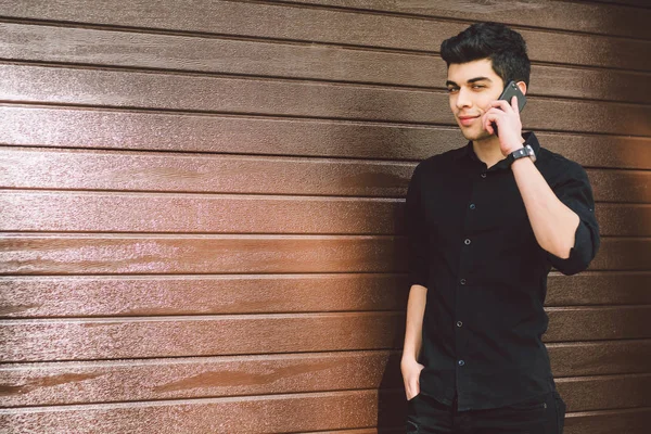 Portrait model young handsome male suspect Turkish middle eastern brunette in black shirt uses hand phone to call technology on the street in sunny weather against the background of a wooden wall