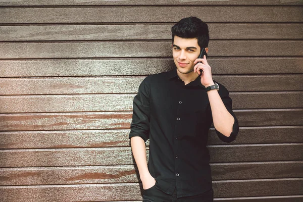 Portrait model young handsome male suspect Turkish middle eastern brunette in black shirt uses hand phone to call technology on the street in sunny weather against the background of a wooden wall