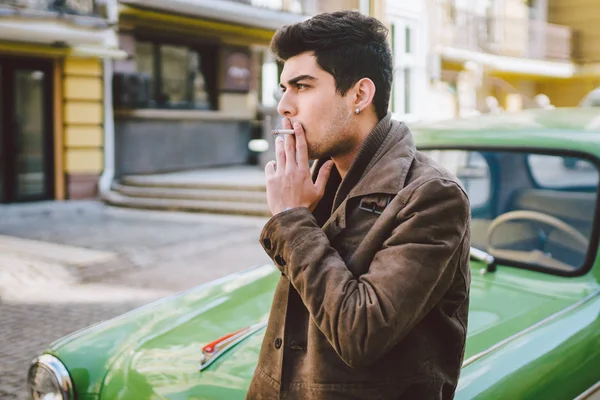Retrato de um modelo jovem bonito sexy masculino morena cara com pele escura turco Médio Oriente morena posando fumar um cigarro perto de carro retro velho na rua — Fotografia de Stock
