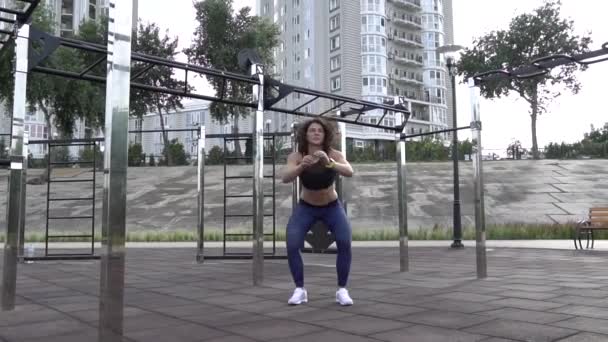 Tema deporte y estilo de vida saludable. Mujer joven caucásica con el pelo rizado tatoo atleta haciendo ejercicio de entrenamiento en los músculos de las piernas y las nalgas. Chica se agacha y salta en el sitio de entrenamiento urbano — Vídeo de stock