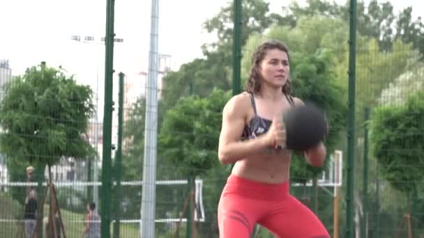 Αθλητισμού και του υγιεινού τρόπου ζωής. Νεαρή γυναίκα Καυκάσιος σγουρά μαλλιά με τατουάζ. Αθλητής κάνει άσκηση κατάρτισης μυς των ποδιών και των γλουτών. Κορίτσι καταλήψεις και άλματα με μπάλα στα χέρια στο γήπεδο πόλη προπόνηση — Αρχείο Βίντεο