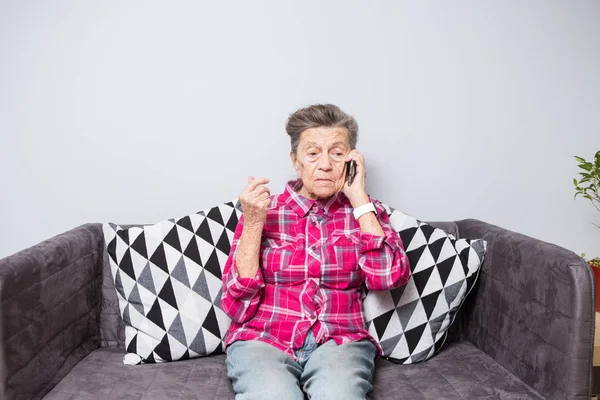 An old elderly woman grandmother with gray hair sits at home on the couch using the hand phone, a telephone conversation to hear the bad news. Emotion fear scare — Stock Photo, Image