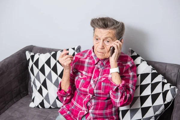 An old elderly woman grandmother with gray hair sits at home on the couch using the hand phone, a telephone conversation to hear the bad news. Emotion fear scare