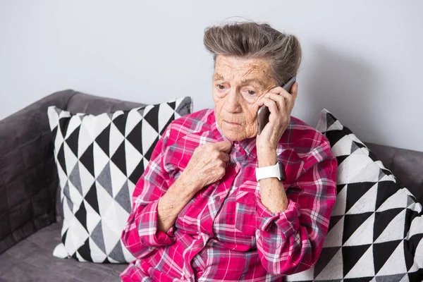 Μια παλιά γιαγιά ηλικιωμένη γυναίκα με γκρίζα μαλλιά στο σπίτι κάθεται στον καναπέ, χρησιμοποιώντας το τηλέφωνο στο χέρι, μια τηλεφωνική συνομιλία για να ακούσει τα άσχημα νέα. Συναίσθημα φόβου τρομάξει — Φωτογραφία Αρχείου