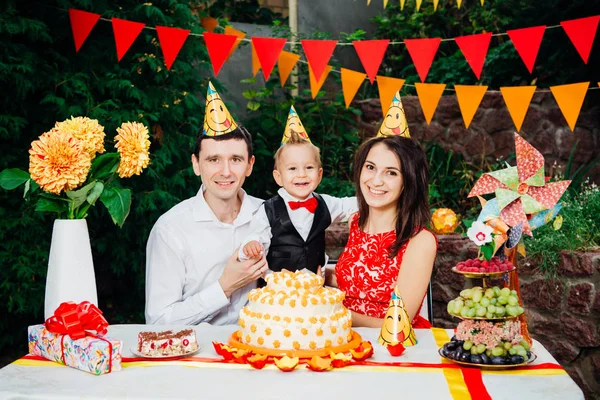 Barn födelsedag tema. familj med tre kaukasiska personer sitter i bakgården av huset på ett festligt dukade bordet i roliga hattar och mössor på huvudet. Glädje och lycka kaka och bakelse mellanmål — Stockfoto