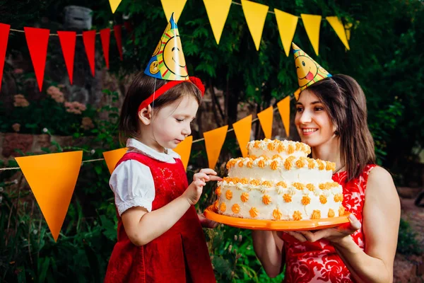 Barnens parti och tårta. Ett litet barn en rolig flicka försöker finger en tårta som mamma nästa håller i händerna. Mot bakgrund av gröna träd och festlig inredning. — Stockfoto