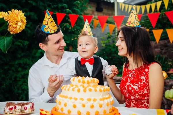 Тема дня рождения детей. Семья из трех кавказцев, сидящих на заднем дворе дома за праздничным украшенным столом в смешных шляпах и кепках на голове. Радость и счастье торт и пирожные — стоковое фото