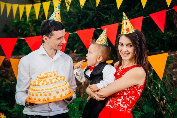 Subject children's birthday party, food and sweets. A young family celebrates one year of son. Dad is holding a big cake, mom is holding a baby in her arms. Baby tastes finger cream on cake with cake