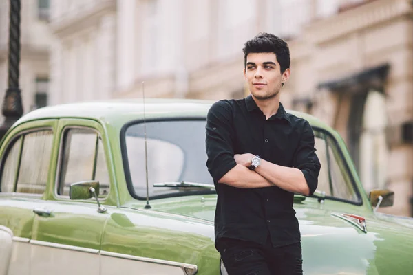 Portrait, handsome, male, model, brunette Mediterranean race Turkish man stands near a retro car of green color posing in a black shirt and jeans in the city — Stock Photo, Image