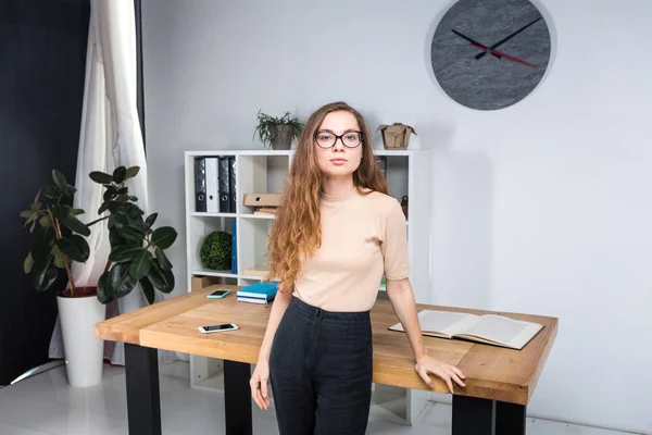 Retrato de uma jovem caucasiana com cabelos longos e óculos para fricção fica em um escritório dentro de uma sala de escritório perto de uma mesa. Uma estudante universitária. Tópico homem aprendizagem — Fotografia de Stock