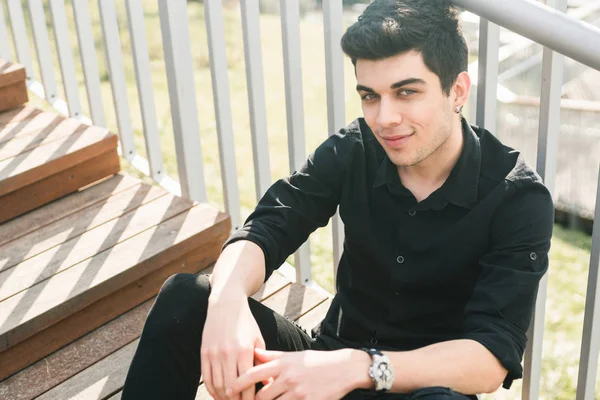 Portrait of a handsome young successful male brunette of a Turkish Mediterranean race sitting on the urban stairs in a black shirt and jeans. Stylish sexy guy with a languid look in sunny weather — Stock Photo, Image