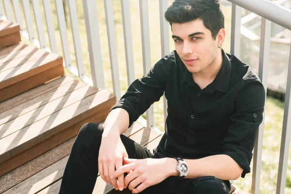 Portrait of a handsome young successful male brunette of a Turkish Mediterranean race sitting on the urban stairs in a black shirt and jeans. Stylish sexy guy with a languid look in sunny weather — Stock Photo, Image