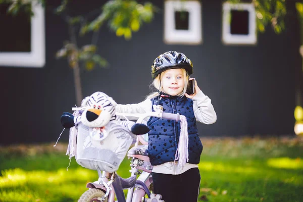 Bambino divertente ragazza caucasica bionda in un casco da bicicletta vicino a una bici viola con un cesto fuori dal parco su un carretto di erba verde a casa. Gioca usa il telefono a mano con sorriso dentato — Foto Stock