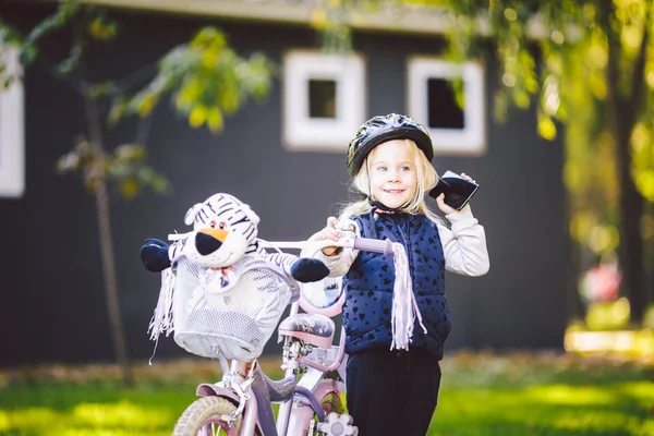 Bambino divertente ragazza caucasica bionda in un casco da bicicletta vicino a una bici viola con un cesto fuori dal parco su un carretto di erba verde a casa. Gioca usa il telefono a mano con sorriso dentato — Foto Stock