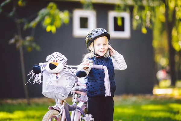 Bambino divertente ragazza caucasica bionda in un casco da bicicletta vicino a una bici viola con un cesto fuori dal parco su un carretto di erba verde a casa. Gioca usa il telefono a mano con sorriso dentato — Foto Stock