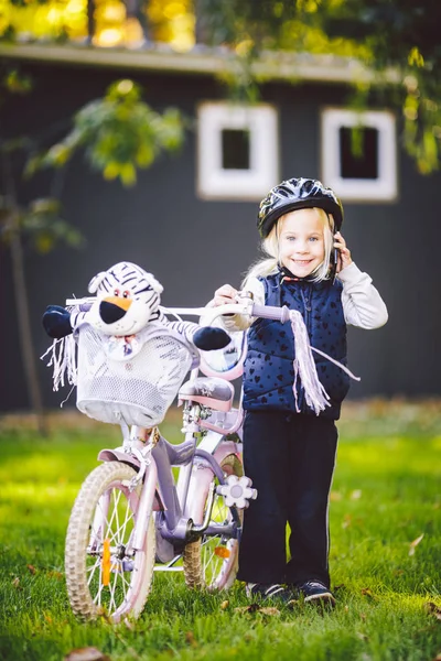 Lustiges kaukasisches Mädchen mit Fahrradhelm in der Nähe eines lila Fahrrads mit einem Korb außerhalb des Parks auf einem grünen Rasengraswagen zu Hause. spielt mit dem Handtelefon mit zahmem Lächeln — Stockfoto