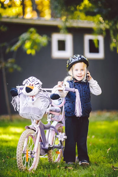 Bambino divertente ragazza caucasica bionda in un casco da bicicletta vicino a una bici viola con un cesto fuori dal parco su un carretto di erba verde a casa. Gioca usa il telefono a mano con sorriso dentato — Foto Stock