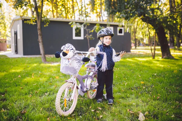 Lustiges kaukasisches Mädchen mit Fahrradhelm in der Nähe eines lila Fahrrads mit einem Korb außerhalb des Parks auf einem grünen Rasengraswagen zu Hause. spielt mit dem Handtelefon mit zahmem Lächeln — Stockfoto