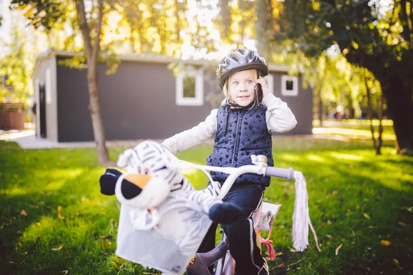Забавная белая девочка-белокурая в велосипедном шлеме рядом с фиолетовым велосипедом с корзиной за пределами парка на зеленой травяной тележке дома. Пьесы используют ручной телефон с зубастой улыбкой — стоковое фото