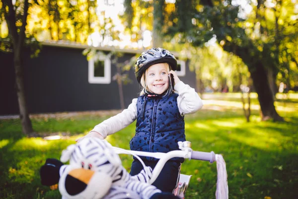 Bambino divertente ragazza caucasica bionda in un casco da bicicletta vicino a una bici viola con un cesto fuori dal parco su un carretto di erba verde a casa. Gioca usa il telefono a mano con sorriso dentato — Foto Stock