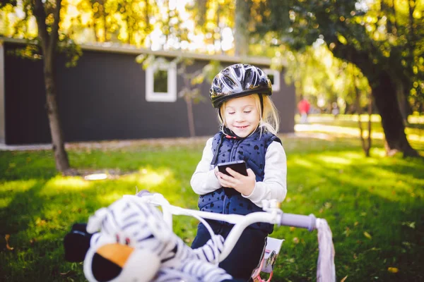 Забавная белая девочка-белокурая в велосипедном шлеме рядом с фиолетовым велосипедом с корзиной за пределами парка на зеленой травяной тележке дома. Пьесы используют ручной телефон с зубастой улыбкой — стоковое фото