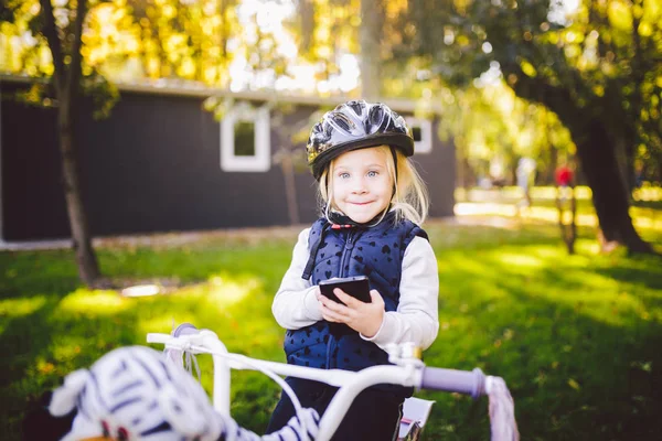 Lustiges kaukasisches Mädchen mit Fahrradhelm in der Nähe eines lila Fahrrads mit einem Korb außerhalb des Parks auf einem grünen Rasengraswagen zu Hause. spielt mit dem Handtelefon mit zahmem Lächeln — Stockfoto