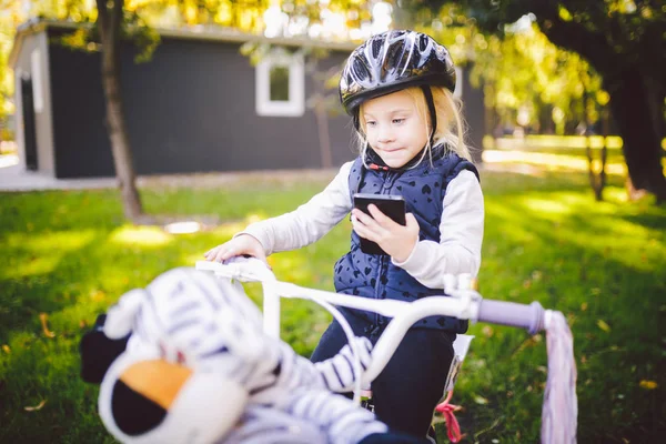 Смішні дитини кавказьких блондинка дівчина в велосипедний шолом поблизу purple велосипед з кошиком в за межами парку на зеленій галявині кошик травою на дому. П'єси використовує рукою телефон з зубастий посмішка — стокове фото