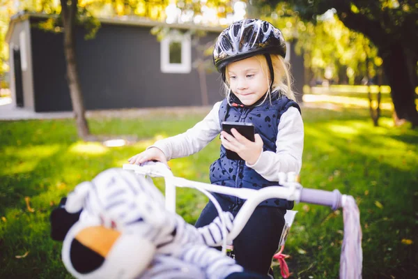 Niño divertido Caucásico chica rubia en un casco de bicicleta cerca de una bicicleta púrpura con una cesta en el exterior del parque en un carrito de césped verde en casa. Juega utiliza teléfono de mano con sonrisa dentada —  Fotos de Stock