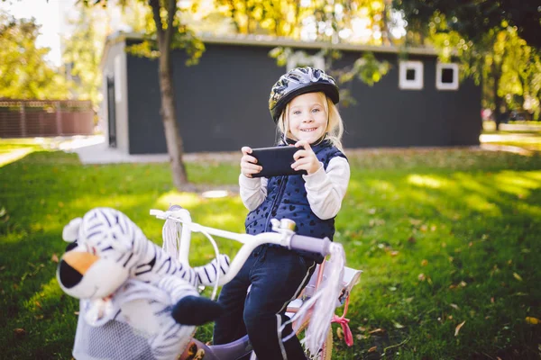 Bambino divertente ragazza caucasica bionda in un casco da bicicletta vicino a una bici viola con un cesto fuori dal parco su un carretto di erba verde a casa. Gioca usa il telefono a mano con sorriso dentato — Foto Stock