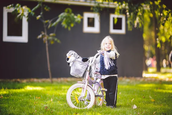 Bambino divertente ragazza caucasica bionda vicino a una bici viola con un cesto e un giocattolo di zebra in un parco esterno su un prato verde erba carrello a casa — Foto Stock