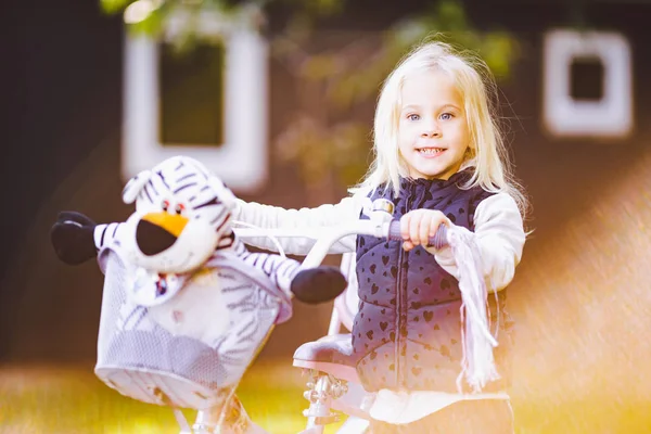 Bambino divertente ragazza caucasica bionda vicino a una bici viola con un cesto e un giocattolo di zebra in un parco esterno su un prato verde erba carrello a casa — Foto Stock