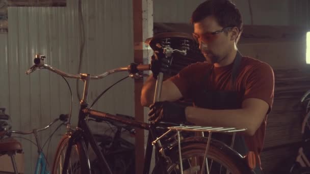 主题小企业自行车维修。一名年轻的白种人黑发男子戴着安全护目镜、手套和围裙, 在车间车库使用手工具修理和调整自行车 — 图库视频影像