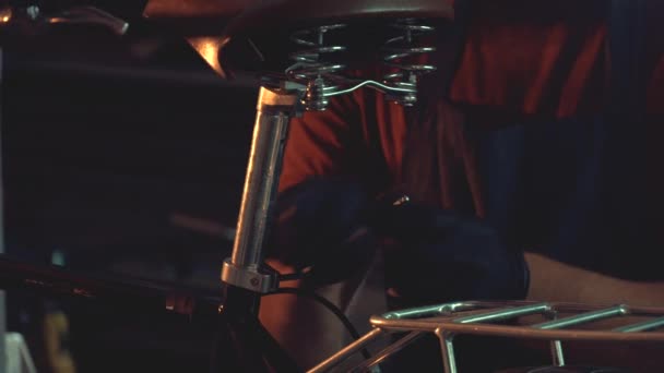 Tema reparação de bicicleta de pequeno negócio. Um jovem homem morena caucasiano usando óculos de segurança, luvas e um avental usa uma ferramenta manual para reparar e ajustar a bicicleta na oficina de garagem — Vídeo de Stock