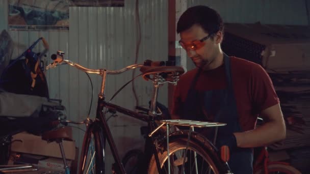 Tema pequeña empresa de reparación de bicicletas. Un joven caucásico morena hombre con gafas de seguridad, guantes y un delantal utiliza una herramienta de mano para reparar y ajustar la bicicleta en el garaje del taller — Vídeo de stock