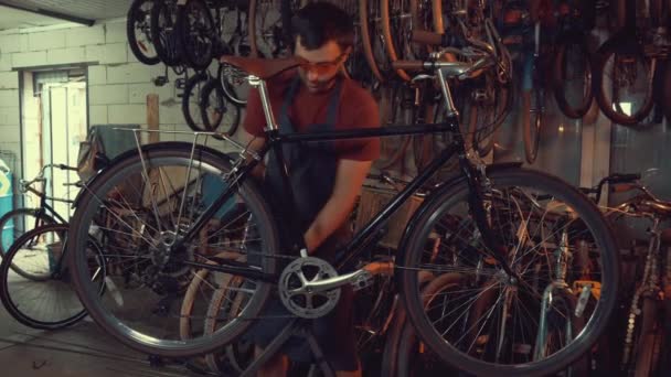 テーマ中小企業の自転車の修理。若い白人ブルネット男安全を身に着けているゴーグル、手袋、エプロンを使用して、手のひらツールを修復し、クランクとペダル システム自転車ガレージ ワーク ショップで調整 — ストック動画